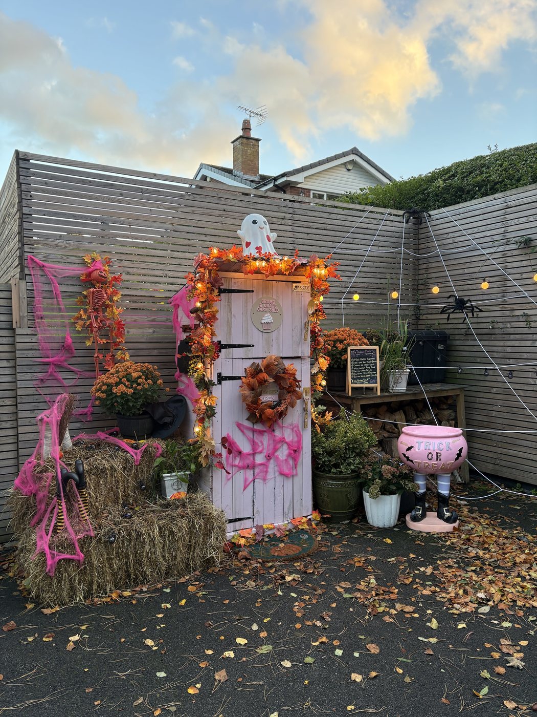 The pink Crumb & Chaos Cake Shed dressed for Halloween with autumn garland, pink cobwebs, hay bales and a trick or treat cauldron