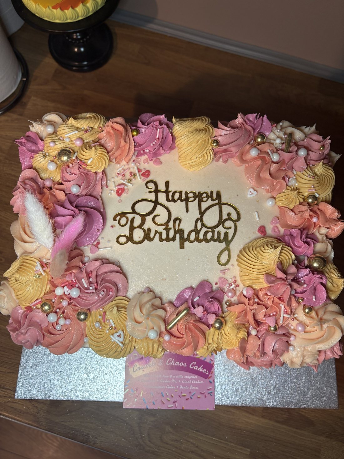 Rectangular birthday sheet cake bordered with pink, coral and yellow buttercream rosettes, pearls and a gold Happy Birthday topper
